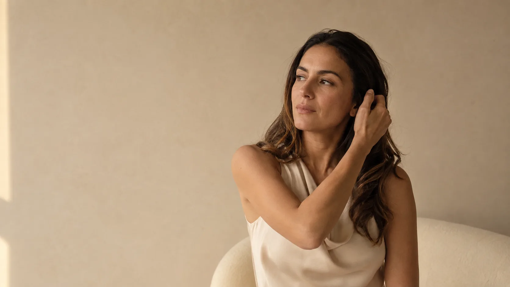 A woman in a sleeveless cream top raises one arm to tuck her hair behind her ear, at ease in soft daylight