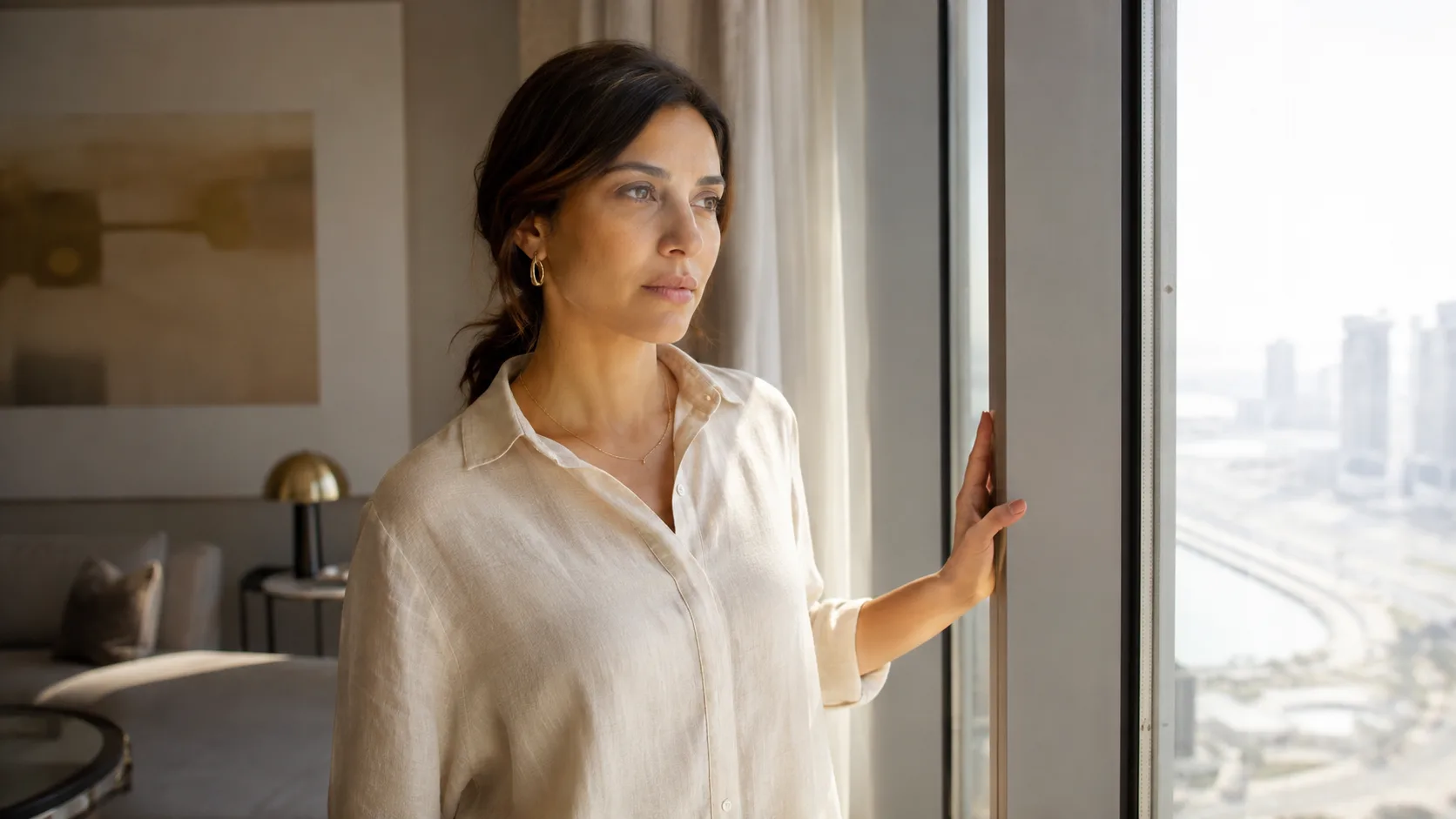 A woman stands at a tall apartment window in Dubai, one hand resting on the frame, looking out at the bright daylight
