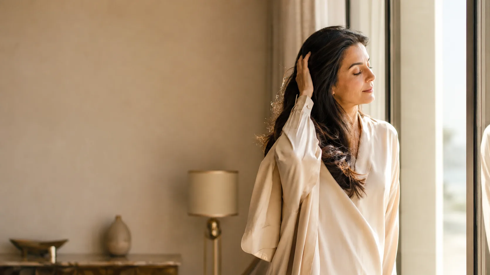 A woman stands at a window running her fingers back through her hair, eyes closed in the warm light
